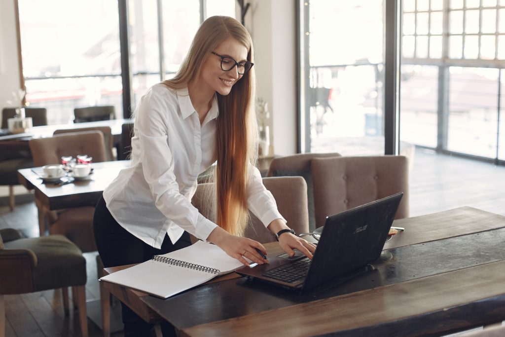 A woman stands at a table, leaning over to type on a laptop.