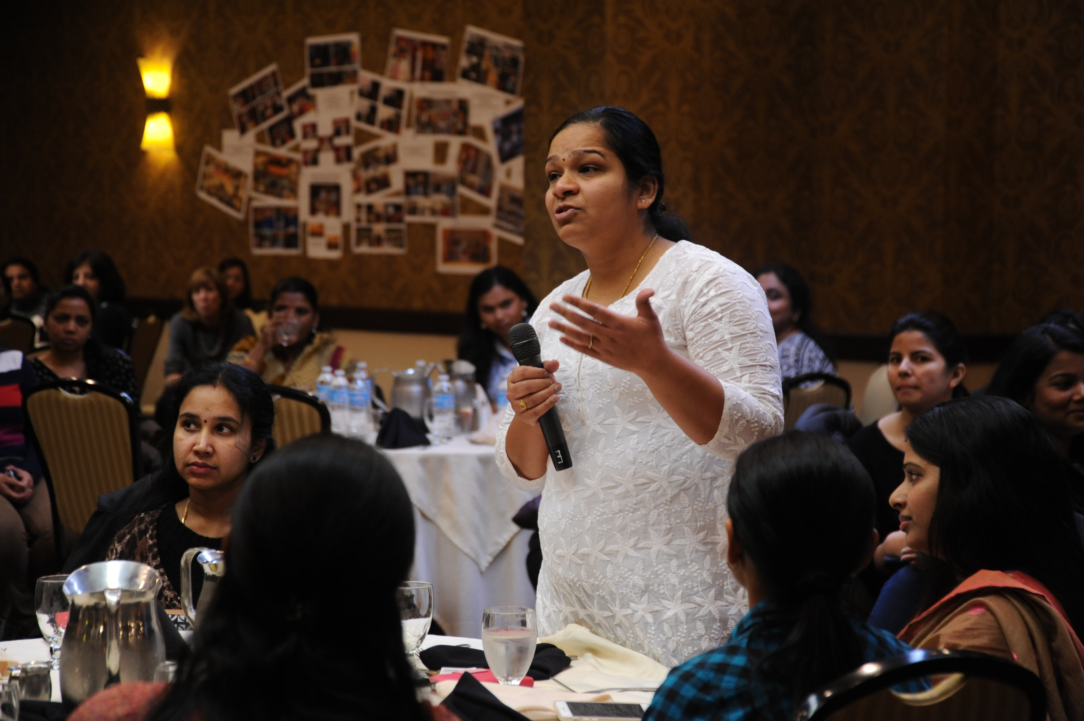 A woman holding a microphone stands up at her table to speak at an event.