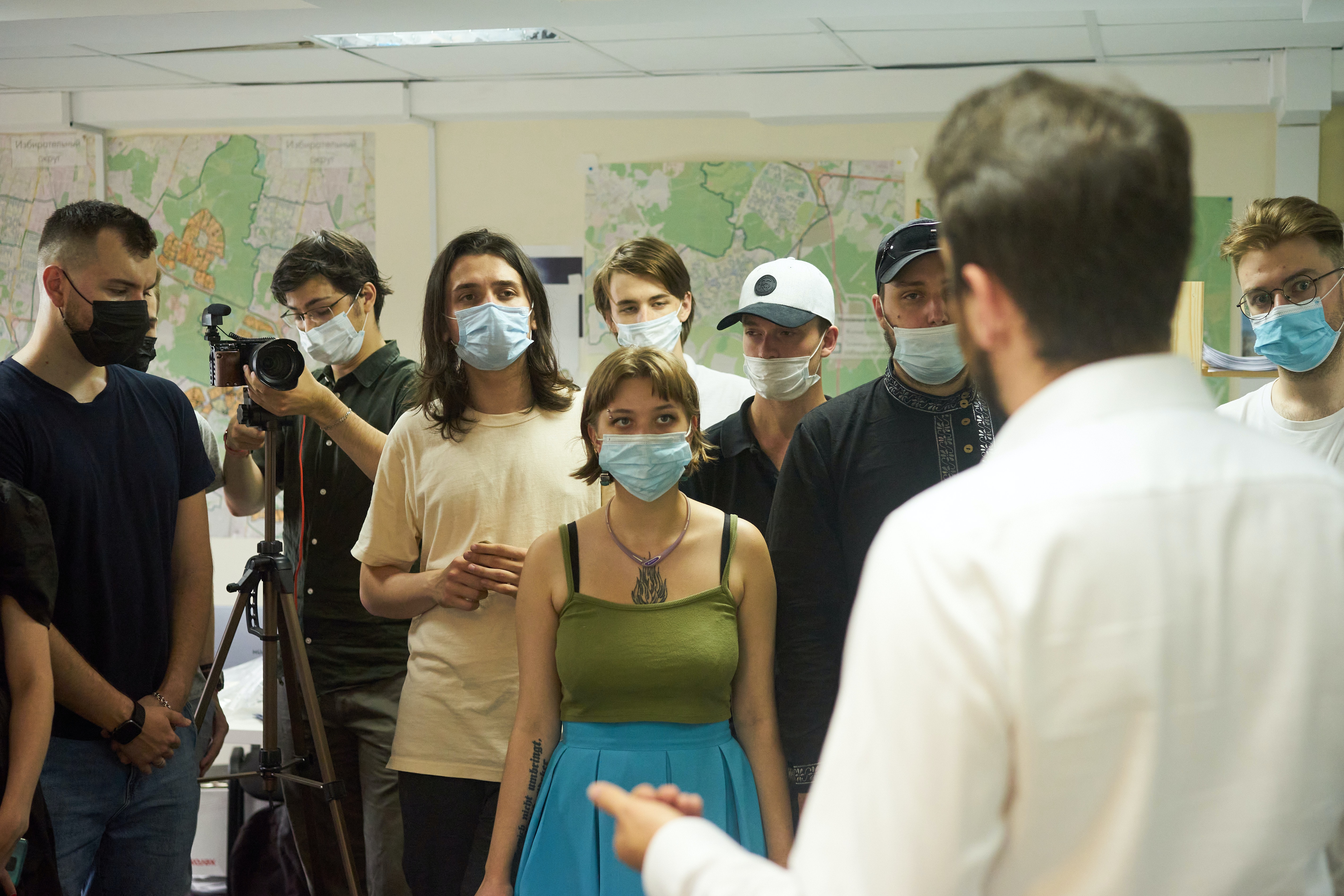 A group of people wearing masks stand in front of a person who is presenting. The room has large maps on the walls.