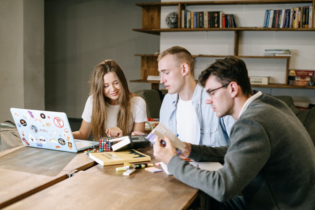 Three people sit together at a table with a laptop open and books on the table and on a bookshelf in the background