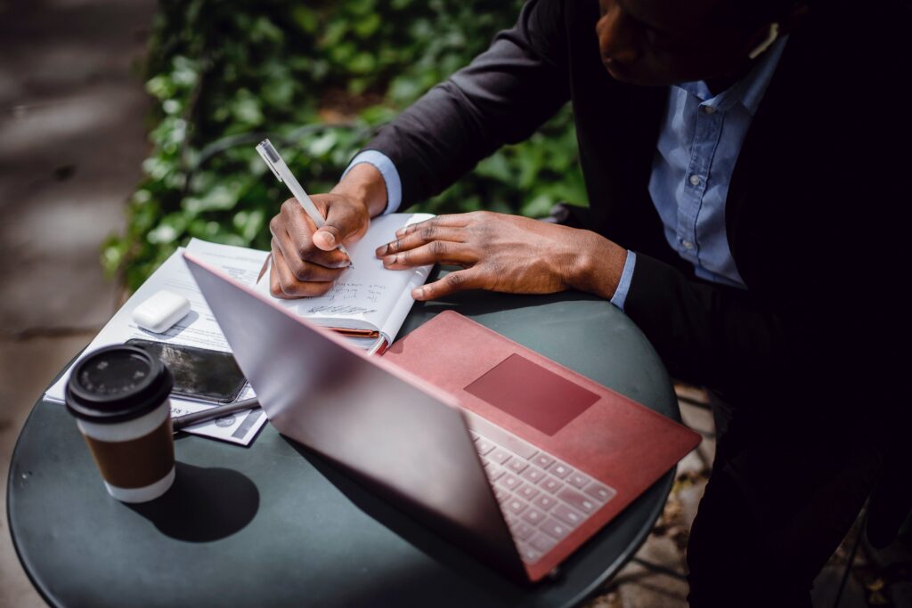 Person writing a plan while sitting at a round table with a notepad, laptop, and coffee.