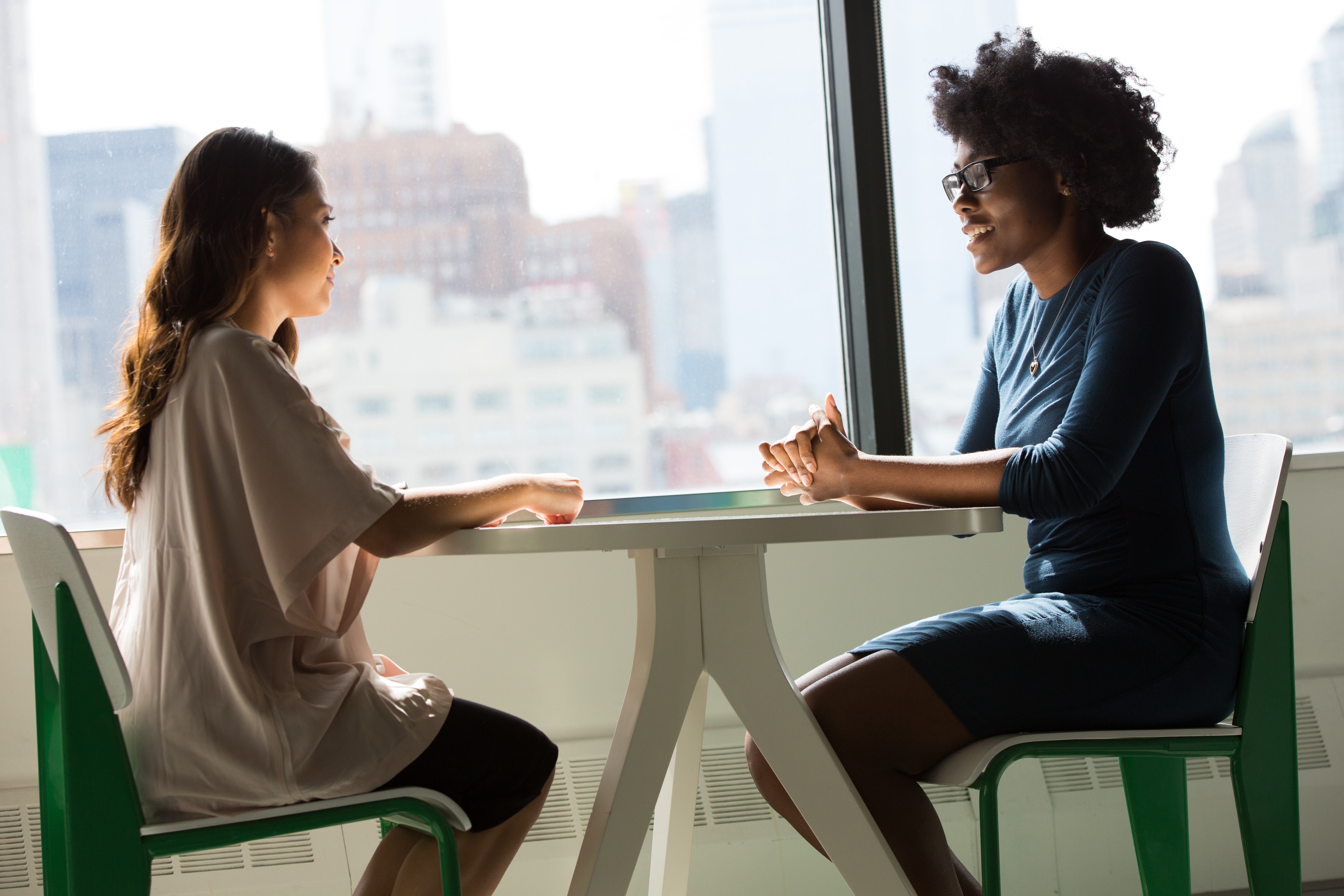 Two women sit at a table inside a tall building, next to a large window with city views.