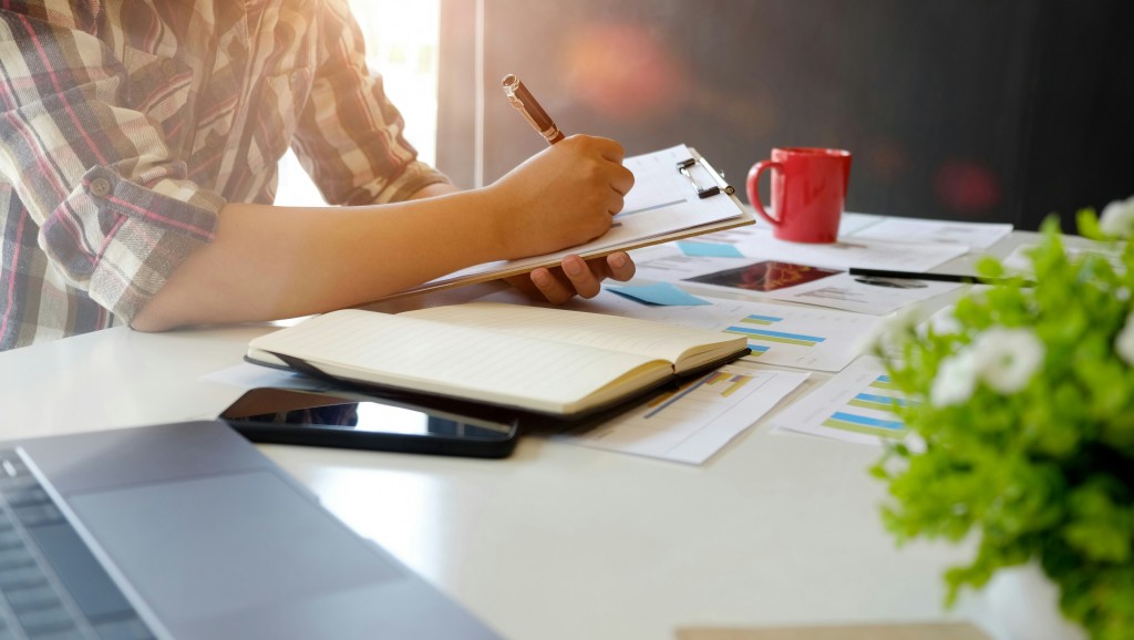 Person writing on a clipboard with documents spread out around them on a desk.