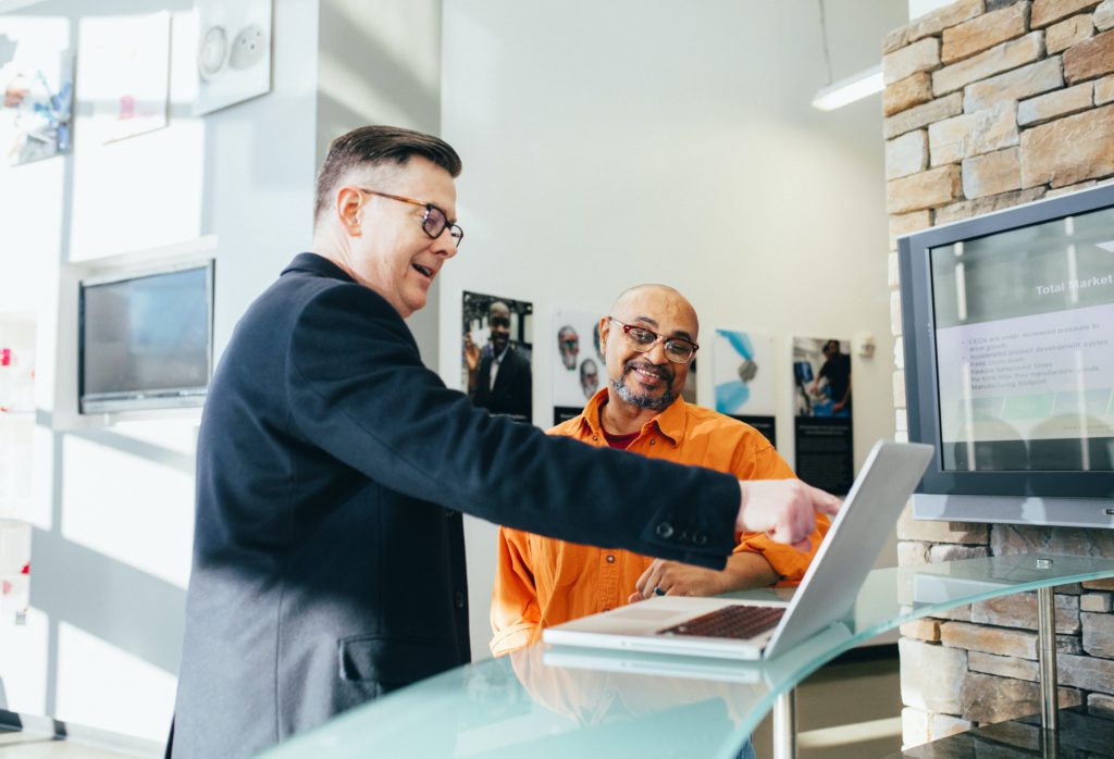Two men in an corporate setting, with both smiling and one pointing at a laptop screen.
